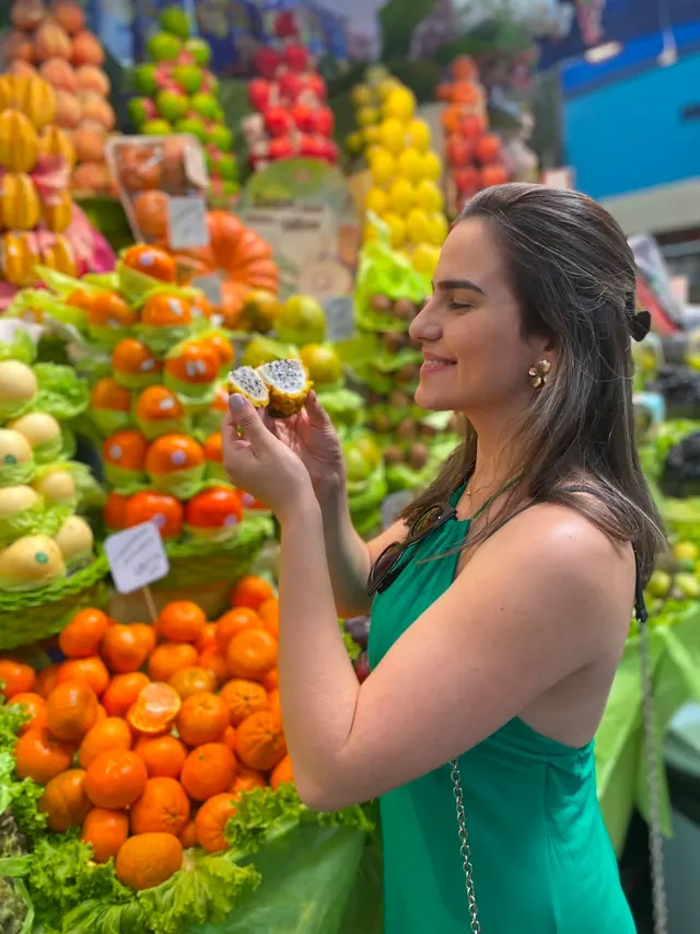 Raissa de vestido verde provando uma pitaia diante de uma banca de frutas