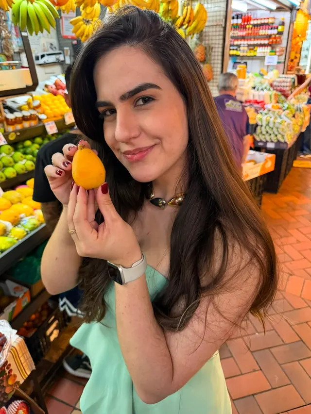 Raissa no mercado, segurando uma nêspera diante das bancas de frutas