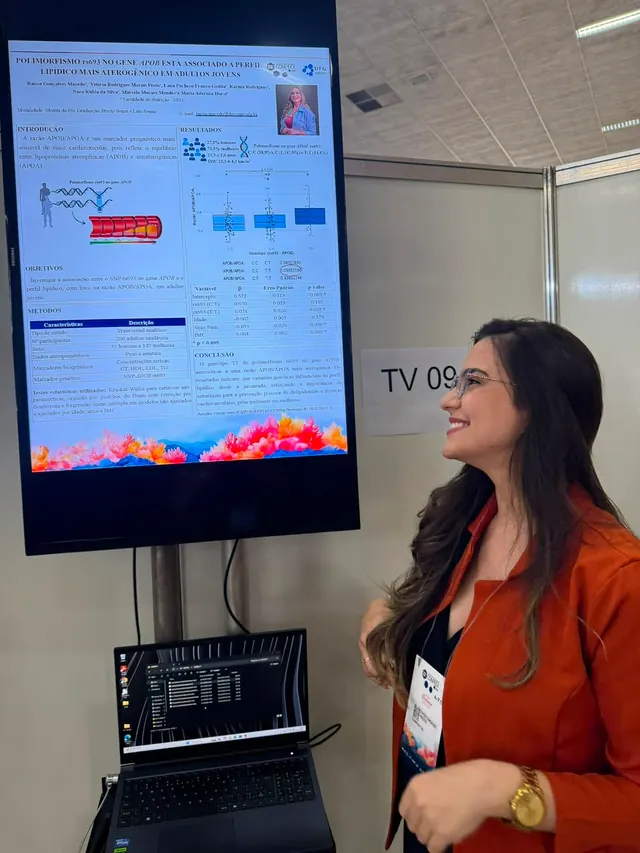 Raissa ao lado da tela onde apresenta um trabalho científico em formato de pôster digital